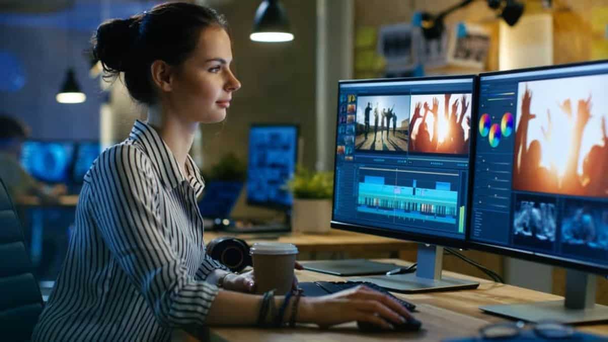 Woman holding a cup of coffee and mouse in front of a dual monitor showing a music video edits on screen.