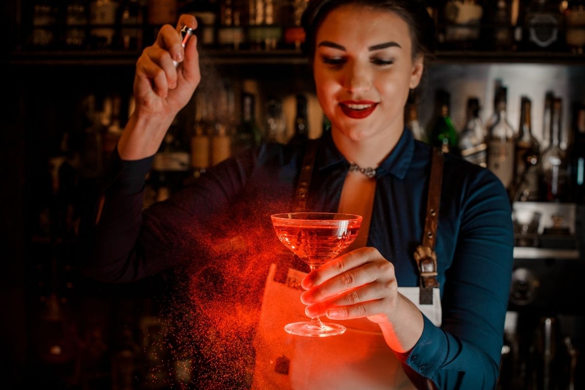 Beautiful bartender girl in the white apron spraying on a delicious cocktail.