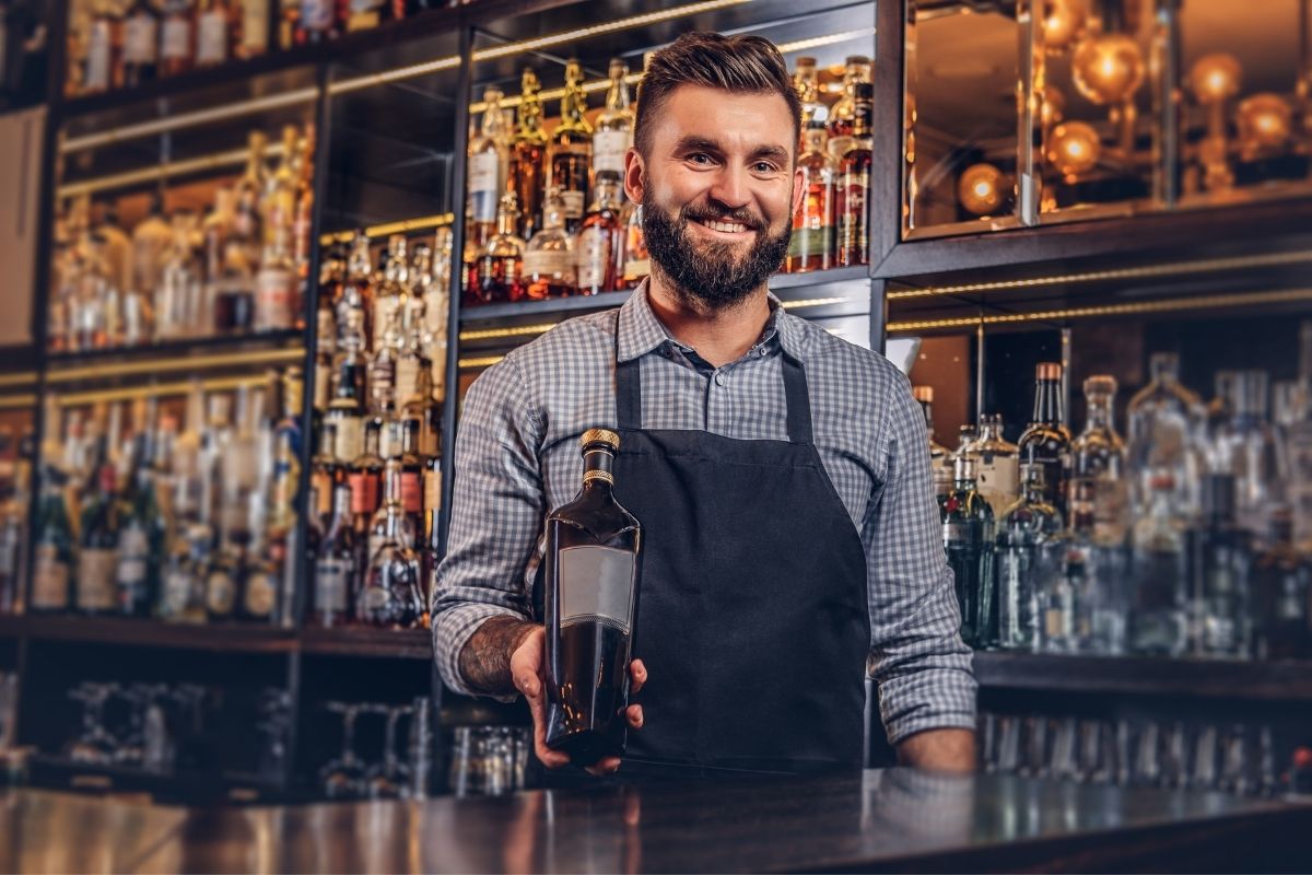 Bartender in a shirt and apron presents a bottle of exclusive alcohol at bar counter.