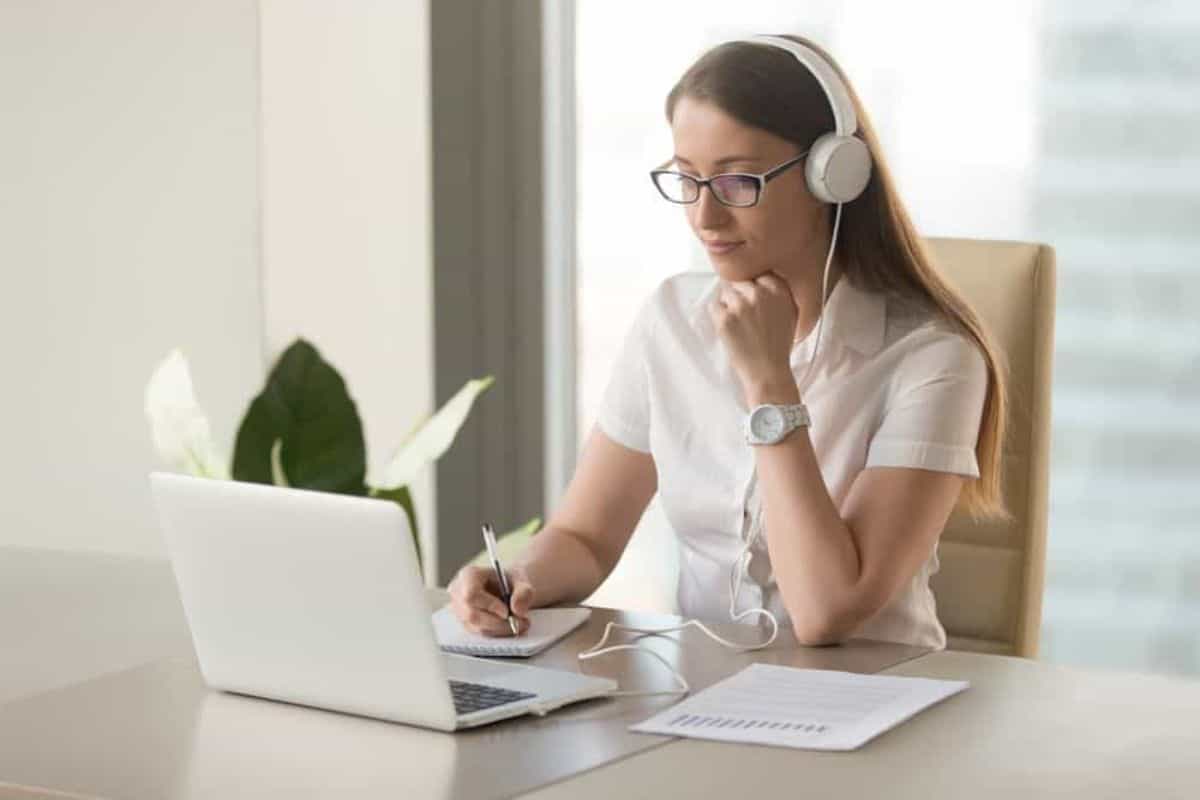 Young woman wearing a headphone and holding a pen on a notebook while sitting in front of a laptop.