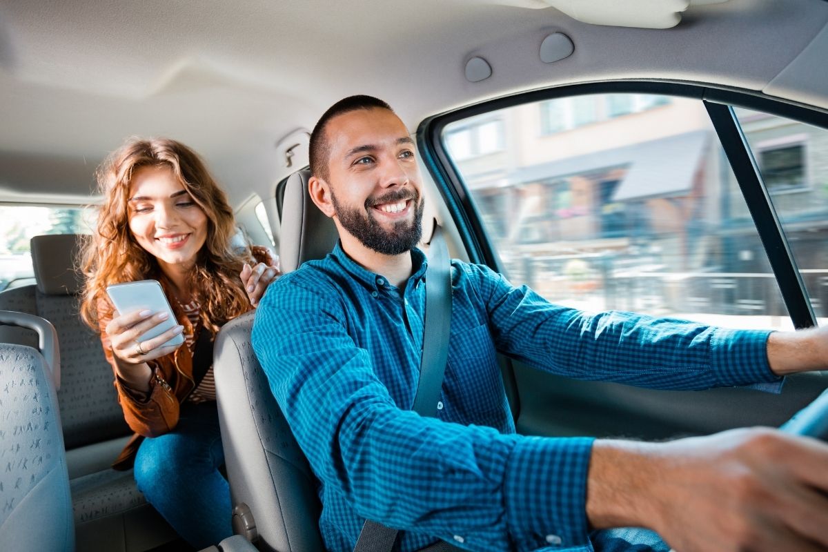 Smiling driver talking with a female passenger while holding a mobile phone.